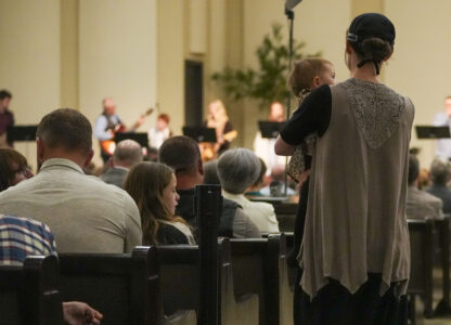 A woman wearing traditional clothing seen in the Mennonite community carries a child while taking a seat during a service at Community Church of Seminole, Sunday, Feb. 23, 2025, in Seminole, Texas. (AP Photo/Julio Cortez)
